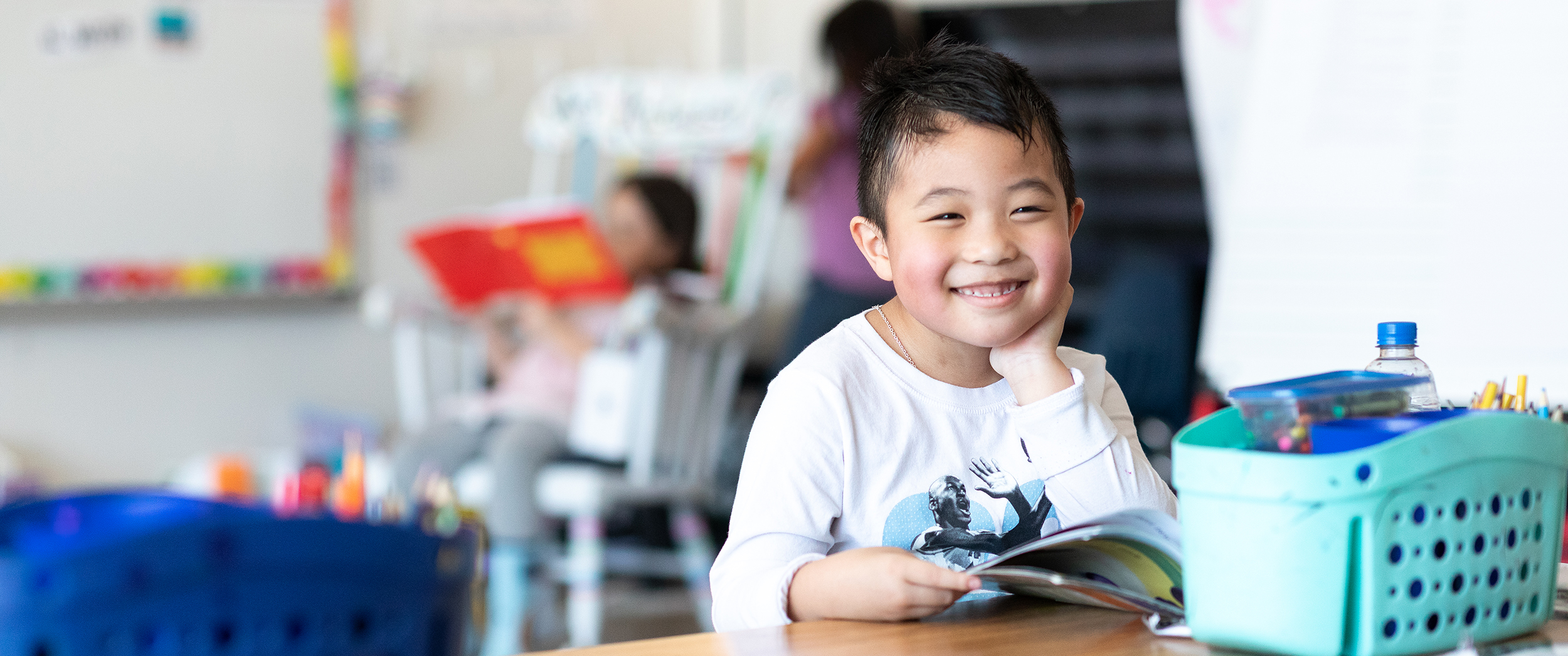 A student sitting at a desk with an opened picture book smiling at the camera